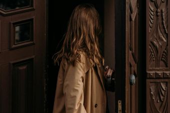 Woman in a brown coat entering through a classic ornate wooden door, creating a fashion narrative.