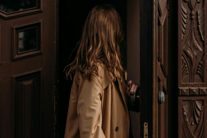 Woman in a brown coat entering through a classic ornate wooden door, creating a fashion narrative.